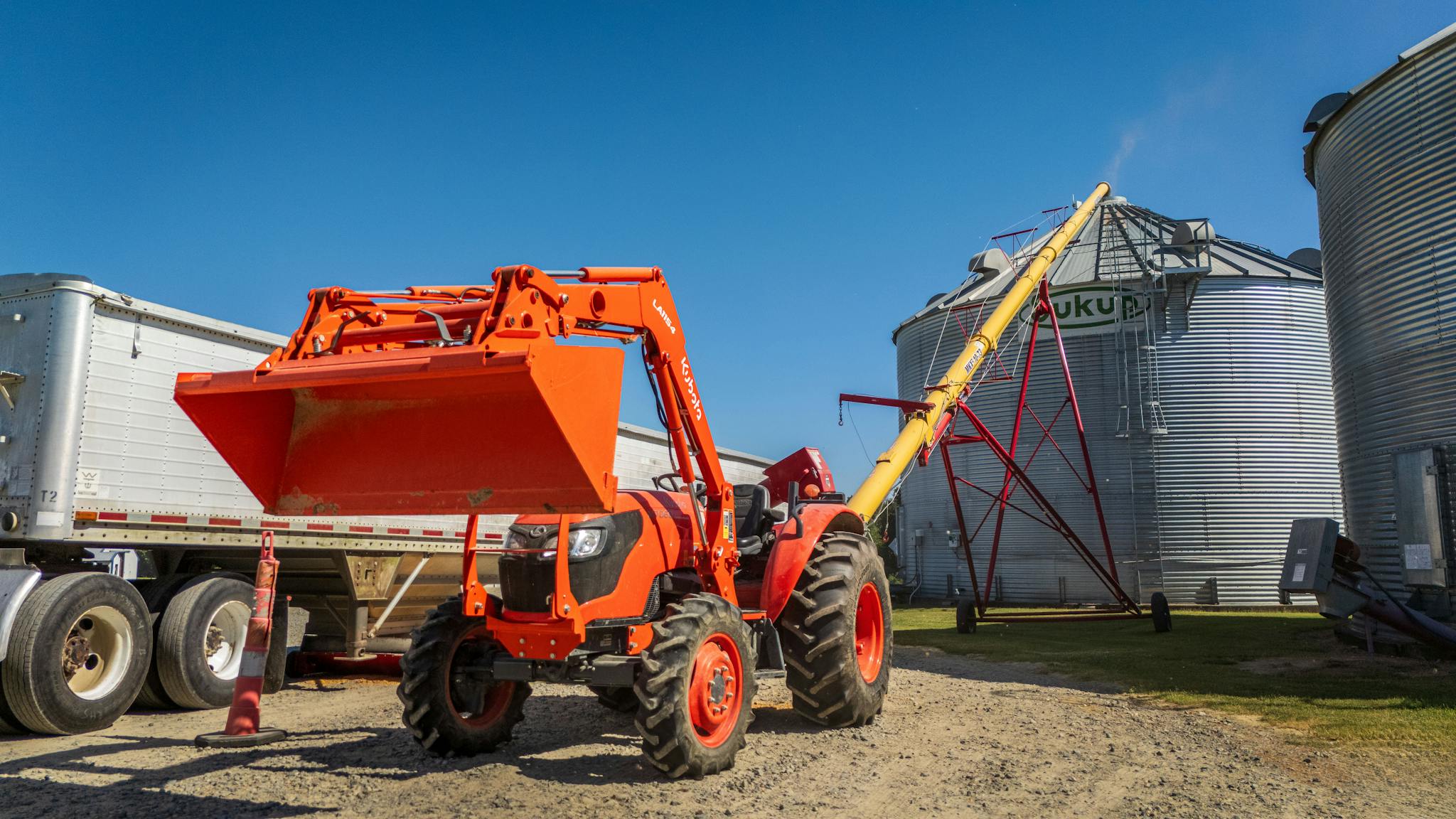 Bright orange tractor with front loader and grain auger near silos under clear sky.